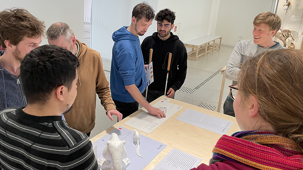 The photo shows students and employees of the Natural History Museum. In front of them on a table are tactile 3D models of a mouse and information boards in Braille and blackletter.