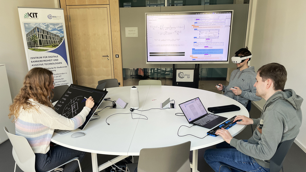 Gruppe von drei Studierenden arbeitet mit unterschiedlichen Hilsmitteln; im Hintergrund ist ein großer Bildschirm mit mathematischen Formeln zu sehen..